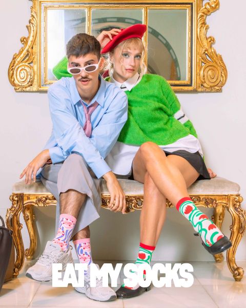 Un giovane uomo e una giovane donna sono seduti su una panchina decorata. Entrambi indossano calzini stravaganti con motivi colorati. L'uomo indossa occhiali da sole e una camicia blu, mentre la donna indossa un basco rosso e un maglione verde. In basso si legge la frase "EAT MY SOCKS".