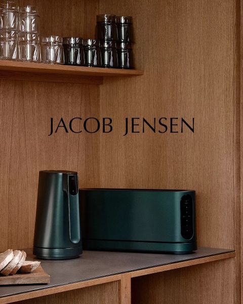 Una cucina moderna con scaffali in legno, bicchieri, una pagnotta di pane e elettrodomestici Jacob Jensen verde scuro sul bancone. Il marchio "JACOB JENSEN" è presente sulla parete a pannelli di legno.
