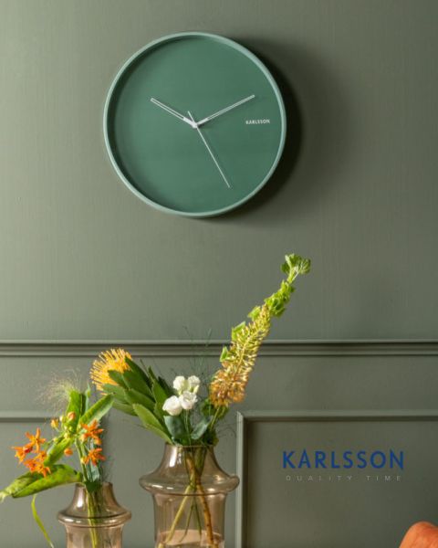 Un orologio da parete minimalista verde con lancette bianche è appeso a una parete verde sopra due vasi di vetro con vari fiori arancioni, bianchi e gialli. Nell'angolo in basso a destra appare in blu la parola "KARLSSON".