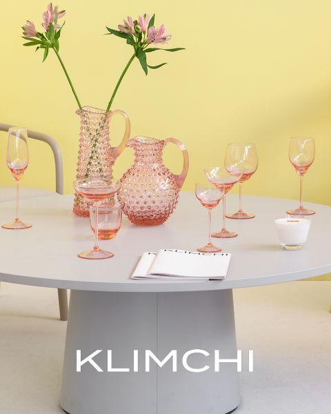 Una tavola elegantemente apparecchiata presenta stoviglie in vetro rosa, tra cui brocche, bicchieri e calici. Fiori rosa in un vaso aggiungono un tocco di eleganza. Lo sfondo è in un tenue giallo e nella parte inferiore dell'immagine si legge la parola "KLIMCHI".