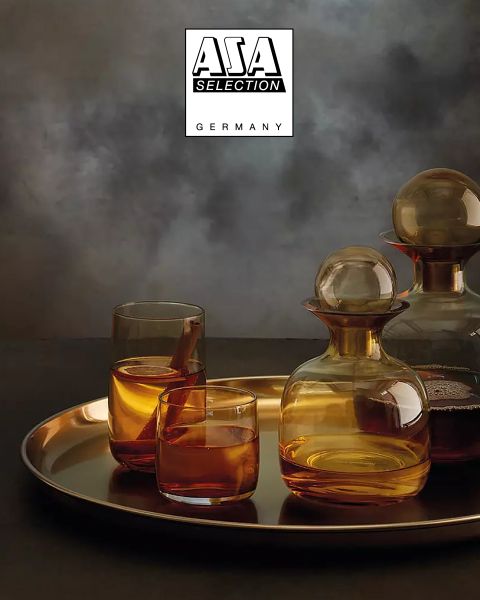 Caraffe color ambra con bicchieri abbinati, riempite con un liquido marrone, sono posizionate su un vassoio dorato davanti a uno sfondo suggestivo con texture grigia. In alto è raffigurato il logo di ASA Selection Germany.