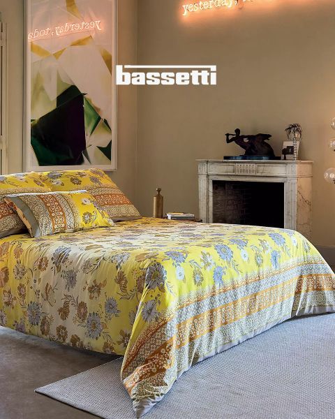 Una camera da letto con un copriletto a fiori gialli, cuscini abbinati, un camino in marmo bianco, arte moderna sulla parete e un'insegna al neon sopra con la scritta "Ieri, oggi". La parola "Bassetti" è ben visibile.