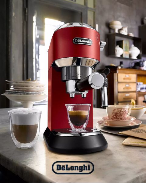 Una macchina da espresso De’Longhi rossa su un bancone della cucina versa il caffè in una tazza di vetro. Sullo sfondo si vedono una tazza di cappuccino, una pila di pancake e una cucina accogliente.