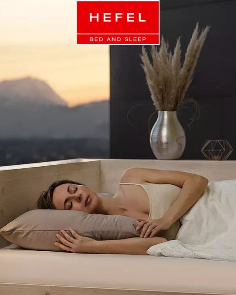 Una donna dorme serenamente su un fianco in un letto moderno con cuscino beige e coperta bianca. Sullo sfondo si vedono un vaso con erba di pampa e una vista sulle montagne al tramonto. In alto campeggia il logo di "Hefel Bed and Sleep".