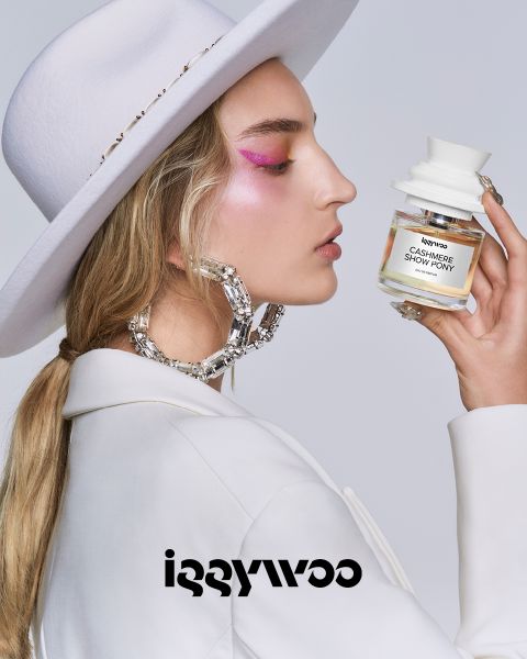 Una persona con i capelli lunghi, un cappello bianco e trucco occhi rosa tiene in mano una bottiglia con la scritta "Cashmere Show Pony" di Iggywoo. Indossa grandi orecchini a cerchio lucidi e una giacca bianca su uno sfondo semplice. In basso si vede il logo Iggywoo.