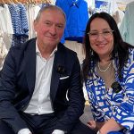Un uomo e una donna siedono sorridendo in un negozio di abbigliamento. Indossano abiti coordinati blu e bianchi. Dietro di loro ci sono appendiabiti con vestiti blu, bianchi e rossi.