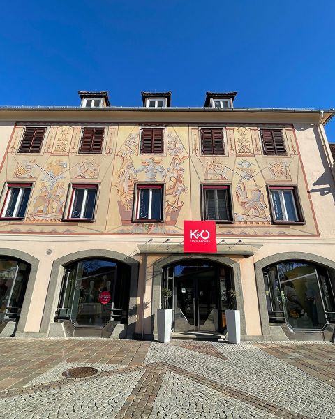 Edificio storico con affreschi elaborati sopra finestre e porte ad arco. Sopra l'ingresso si trova un'insegna rossa e bianca con la scritta "K&U". La facciata colpisce per i motivi artistici e un cielo azzurro brillante sullo sfondo.