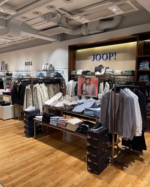 Interno di un negozio di abbigliamento con scaffali pieni di abiti da uomo, camicie e giacche. Sono esposti marchi noti come "Boss" e "Joop!". Capi di abbigliamento ordinatamente piegati sono impilati sugli scaffali, tra cui si possono vedere alcune scarpe. Il pavimento è in legno.