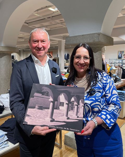Un uomo in giacca e cravatta e una donna con un abito a motivi blu sono in piedi sorridenti in un edificio e tengono in mano un libro. La copertina mostra una scena architettonica con archi. Sullo sfondo si vedono appendiabiti e colonne ad arco.