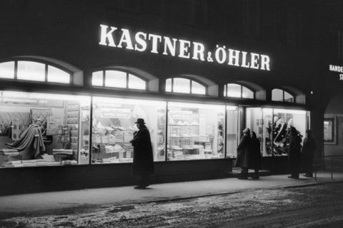 Immagine in bianco e nero di un negozio Kastner & Öhler di notte. Quattro persone in lunghi cappotti e cappelli sono in piedi sulla strada innevata e guardano le vetrine con tessuti piegati e vari articoli. Il negozio è illuminato e crea un contrasto con l'ambiente circostante buio.