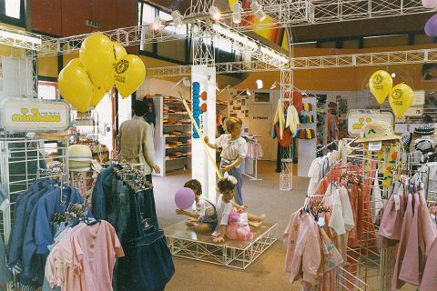 Bambini giocano su un palcoscenico in un negozio di abbigliamento colorato pieno di appendiabiti. Palloncini giallo brillante fluttuano sopra le loro teste e sono in vendita vari capi di abbigliamento colorati e abiti casual.