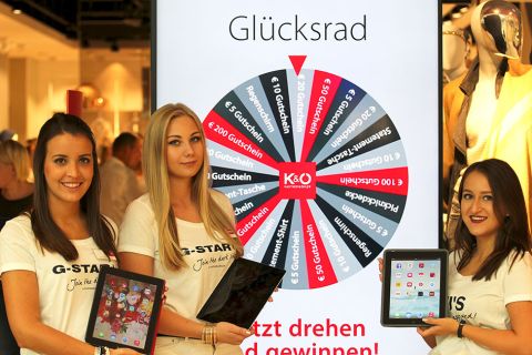 Tre donne in magliette bianche G-STAR tengono dei tablet davanti a un display "Ruota della Fortuna" in un negozio. Sulla ruota si possono vincere diversi premi come buoni e promozioni.