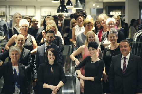 Un folto gruppo di persone vestite in modo professionale posa su una scala mobile in un ambiente interno. Il gruppo è composto da uomini e donne con diverse espressioni facciali. Sullo sfondo si intravede un interno moderno con scaffali e lampade a sospensione.