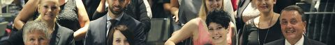 Un folto gruppo di persone vestite in modo professionale posa su una scala mobile in un ambiente interno. Il gruppo è composto da uomini e donne con diverse espressioni facciali. Sullo sfondo si intravede un interno moderno con scaffali e lampade a sospensione.