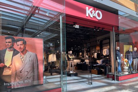 Vetrine di K&Ö, un negozio di abbigliamento con ingresso in vetro. A sinistra nell'immagine, un grande poster con due uomini in abiti alla moda. Nel negozio, manichini presentano vari capi di abbigliamento. Il negozio è arredato in stile moderno e l'abbigliamento è presentato in modo chiaro.