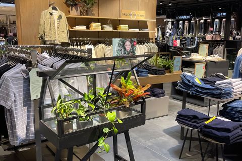 Interni di un negozio di abbigliamento con abbigliamento casual su scaffali e appendiabiti, inclusi jeans e camicie. Una piccola vetrina di vetro con piante in vaso è posizionata centralmente, contribuendo a un'atmosfera naturale ed ecologica.