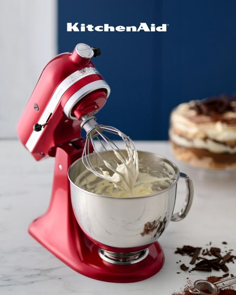 Un robot da cucina KitchenAid rosso con panna montata sulla frusta è posizionato su un piano di marmo. Sullo sfondo si intravedono un dessert a strati e scaglie di cioccolato, sopra c'è scritto "KitchenAid".