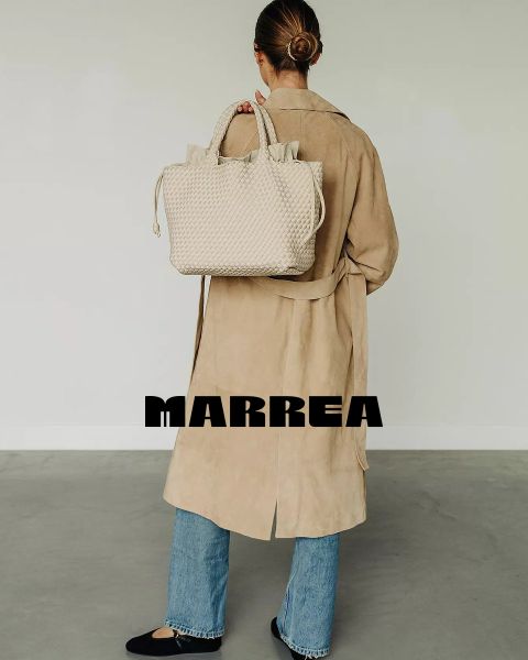 Una donna con uno chignon ordinato, un lungo cappotto beige, blue jeans e scarpe nere è in piedi di spalle e tiene in mano una grande borsa a mano intrecciata color crema. Sulla parte bassa della schiena ha la scritta "MARREA".