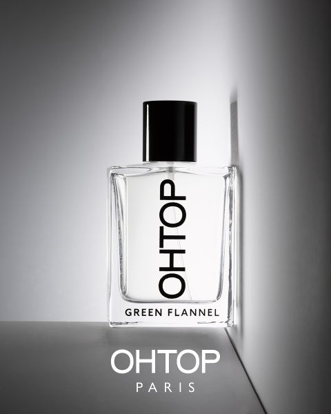Il flacone di profumo con la scritta "OHTOP GREEN FLANNEL" è posizionato su una superficie grigia davanti a uno sfondo grigio chiaro. La bottiglia è parzialmente ombreggiata, sotto c'è la scritta "OHTOP PARIS". Design minimalista con chiusura nera.