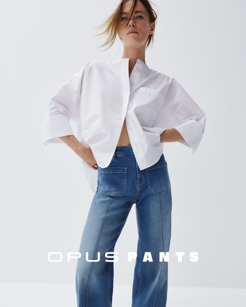 Persona in camicia bianca leggera e jeans, mani sui fianchi. In basso c'è il testo "Opus Pants". I capelli sono leggermente scompigliati dal vento, sfondo bianco.