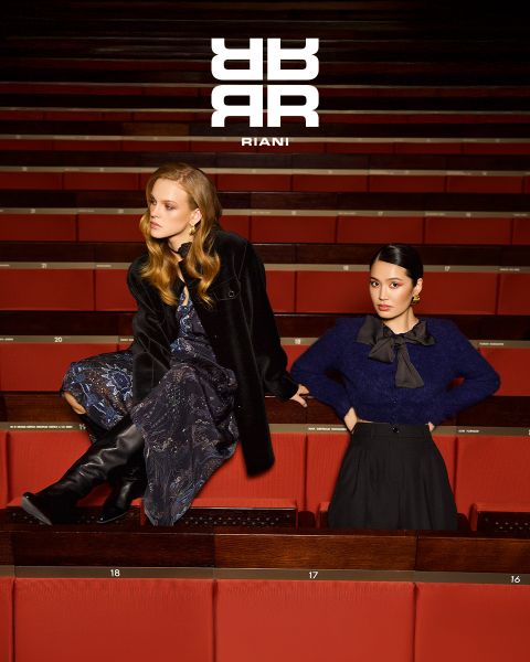 Due donne posano con stile in un teatro con sedili rossi. Una è seduta sullo schienale di un sedile e indossa un abito fantasia, un cappotto nero e stivali. L'altra è in piedi accanto a lei con un maglione blu navy e una gonna nera. Sopra di loro appare il logo "RIANI".
