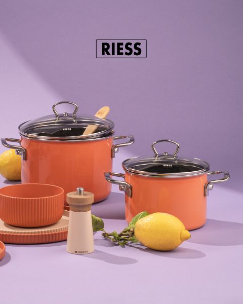 Due pentole arancioni con coperchi in vetro, un cucchiaio di legno e un macinapepe sono su uno sfondo viola. Accanto ci sono un limone, bacche verdi, ciotole arancioni impilate e piatti beige. Sopra si vede il marchio "RIESS".