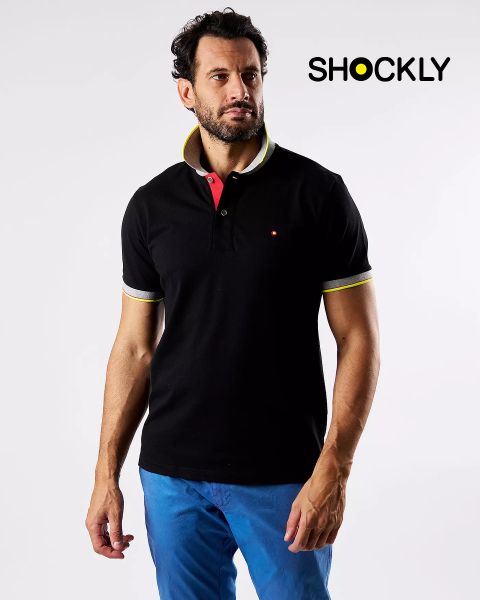 Un uomo con una polo nera con dettagli giallo-bianchi e pantaloni blu è in piedi davanti a uno sfondo bianco semplice. In alto a destra, la parola "SHOCKLY" è scritta in nero.