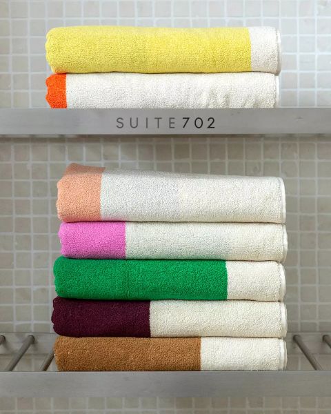 Sei asciugamani piegati ordinatamente con estremità colorate - giallo, arancione, marrone, rosa, verde e bordeaux - sono impilati su due ripiani davanti a una parete piastrellata. Sul ripiano superiore c'è la scritta "SUITE702".