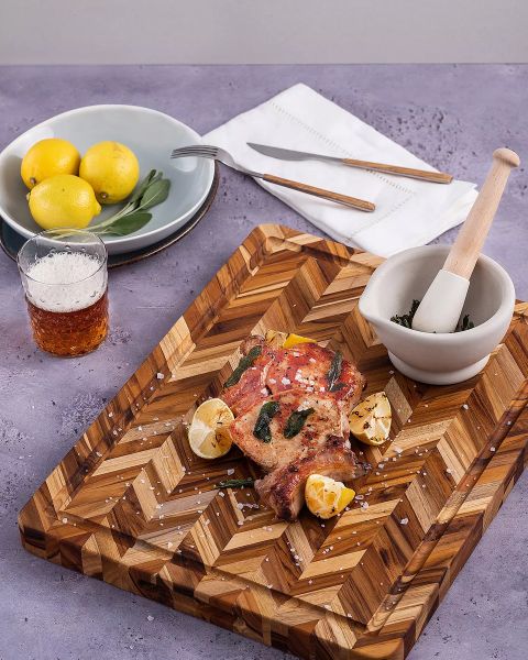Un tagliere di legno con carne speziata e cotta, spicchi di limone e sale grosso. Accanto ci sono un mortaio con erbe aromatiche, un bicchiere con una bevanda, un piatto con limoni, e forchetta e coltello su un tovagliolo, il tutto su una superficie viola.