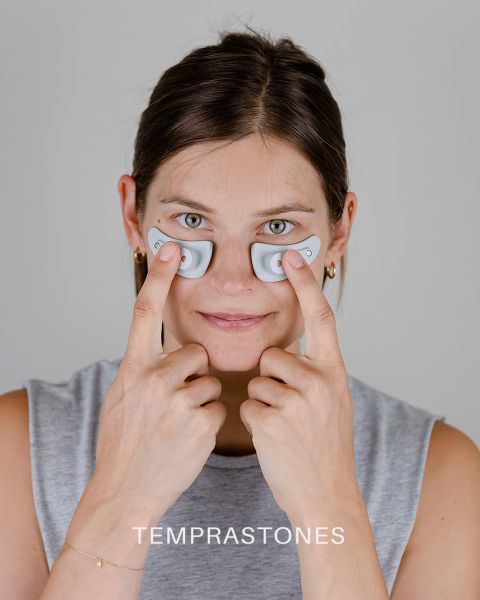 Una persona tiene due piccoli dispositivi ovali sotto gli occhi e li preme delicatamente sulla pelle. Ha un'espressione neutra e indossa un top senza maniche. Nella parte inferiore dell'immagine appare la parola "TEMPRASTONES".
