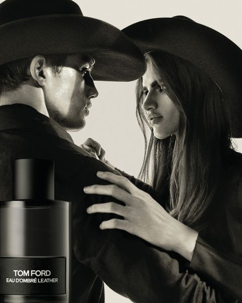 Un uomo e una donna con cappelli a tesa larga si fronteggiano con espressioni tese. In primo piano c'è una bottiglia di Eau d'Ombré Leather di Tom Ford. L'immagine è in bianco e nero e trasmette un'impressione drammatica e classica.