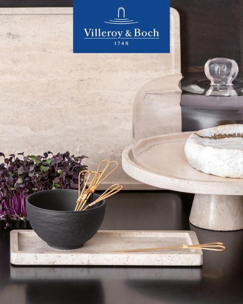 Una mostra di cucine espone una ciotola nera con piccole fruste su un vassoio rettangolare. Accanto si trova un piatto con coperchio di vetro, su cui sono disposti formaggio ed erbe fresche. In alto spicca il logo di Villeroy & Boch.
