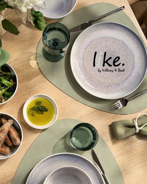 Vista dall'alto di un tavolo da pranzo con un piatto bianco e blu con la scritta "like. by Villeroy & Boch", tovagliette verdi, bicchieri di vetro trasparenti, una ciotola di grissini, una ciotola di insalata e una ciotola di olio, guarnita con erbe aromatiche.