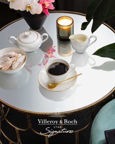 Su un tavolino da caffè in vetro riflettente sono posizionate una tazza di caffè in una tazza bianco-oro, una piccola teiera, una lattiera, una candela e un piatto di biscotti. La scena appare elegante e accogliente, e in basso si vede il logo di Villeroy & Boch.
