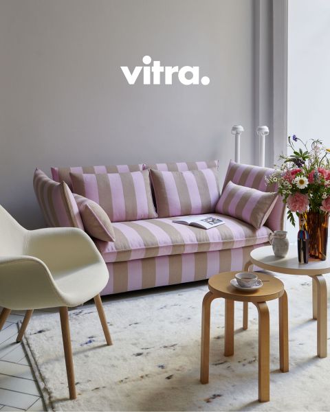 Un soggiorno moderno presenta un divano a righe rosa-beige, una sedia bianca con gambe in legno e un tavolino da caffè rotondo in legno. Sul tavolo c'è un vaso con fiori colorati, sopra c'è scritto "vitra." sulla parete. Il pavimento è ricoperto da un morbido tappeto.
