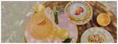 Un tavolo di legno con una brocca di vetro strutturato, un bicchiere rosa, limoni interi con foglie, piatti decorativi impilati, mezza arancia e un piatto di piccoli biscotti rotondi nella luce solare screziata.