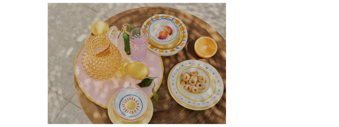 Un tavolo rotondo di legno con piatti colorati, una ciotola di biscotti, arance dimezzate, limoni e cristalleria strutturata, il tutto disposto su una tovaglia a righe rosa nella luce naturale del sole.