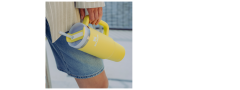 Persona in pantaloncini di jeans tiene un grande bicchiere giallo con manico e cannuccia e indossa una giacca chiara.