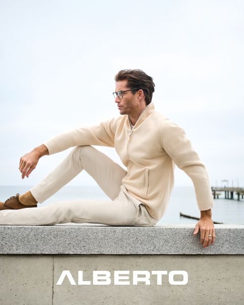 Un uomo con gli occhiali, un maglione beige con la zip e pantaloni chiari è seduto su una sporgenza di pietra in riva al mare. Guarda di lato, sotto di lui c'è scritto "ALBERTO" in bianco. Lo sfondo è nuvoloso e si vede un molo.