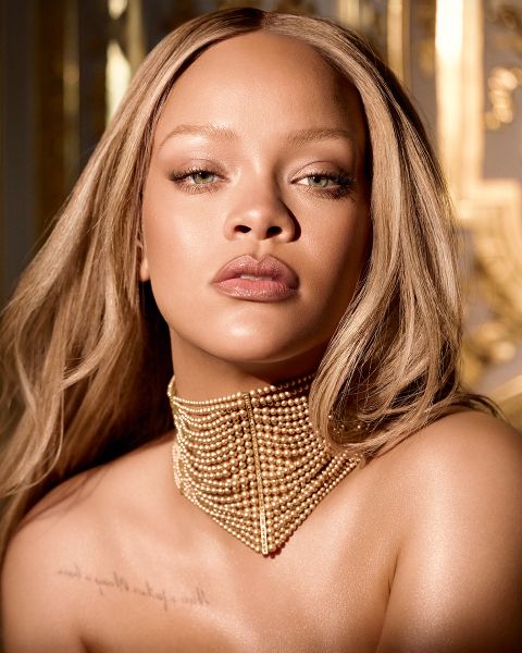 Una donna con lunghi capelli biondi lisci e una collana di perle dorate posa in un ambiente delicatamente illuminato con decorazioni dorate. Indossa un trucco neutro e ha un tatuaggio visibile sulla clavicola.