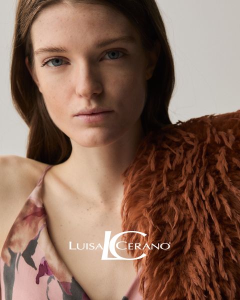 Una donna con lunghi capelli castani indossa un abito a fiori e un capo in pelliccia strutturato color ruggine sulla spalla. Guarda direttamente la fotocamera. In basso al centro si legge il testo "Luisa Cerano".