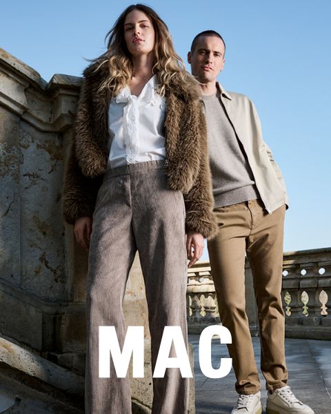 Una donna con una giacca di pelliccia e pantaloni ampi è in piedi accanto a un uomo con una giacca beige e pantaloni color kaki. Entrambi posano con sicurezza all'aperto di fronte a una ringhiera di pietra sotto un cielo blu. La parola "MAC" appare in basso.