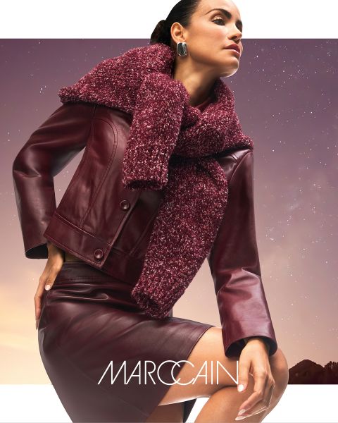 Una donna posa con sicurezza in una giacca di pelle color vinaccia e una gonna con una sciarpa grossa e strutturata color vinaccia. Sullo sfondo si vede un cielo cupo e il marchio "Marc Cain" è visibile in basso.