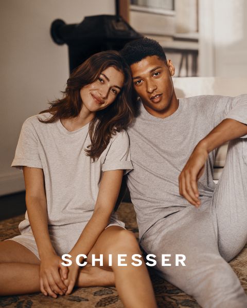 Una donna e un uomo in abbigliamento da casa grigio chiaro siedono vicini su un tappeto e sorridono dolcemente alla fotocamera. Il marchio "SCHIESSER" è visibile nella parte inferiore dell'immagine.