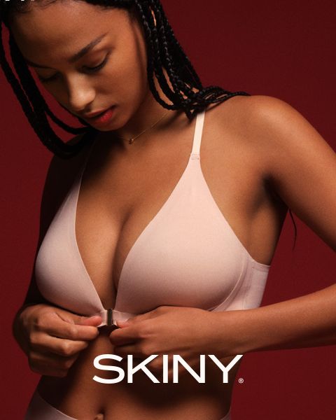Una donna con i capelli intrecciati allaccia un reggiseno rosa chiaro con chiusura frontale su uno sfondo rosso scuro. La parola "SKINY" appare nella parte inferiore dell'immagine.