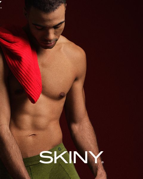 Un uomo muscoloso e alla mano guarda in basso. Ha un maglione rosso sulla spalla e indossa biancheria intima verde. Lo sfondo è rosso scuro e la parola "SKINY" è visualizzata in basso.