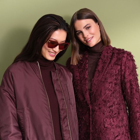 Due donne sono in piedi fianco a fianco davanti a uno sfondo verde chiaro. Una indossa occhiali da sole e una giacca color vinaccia, l'altra un blazer strutturato color vinaccia. Entrambe hanno i capelli scuri e sorridono.