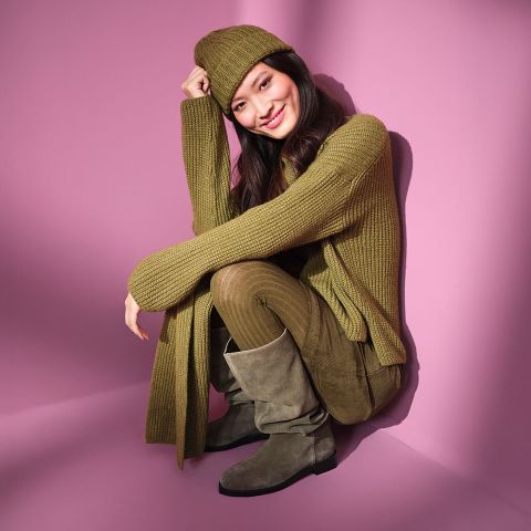 Una donna siede sorridendo di fronte a uno sfondo rosa, indossando un maglione di lana verde oliva, un cappello abbinato, una sciarpa, pantaloni e stivali di camoscio grigi.