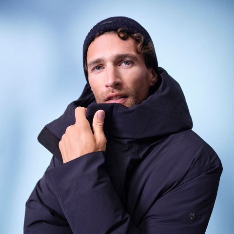 Un uomo che indossa un cappotto invernale scuro e un berretto di lana scuro guarda la telecamera e si tiene il colletto del cappotto davanti al viso su uno sfondo blu tenue.