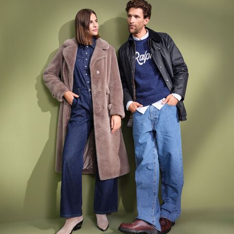 Una donna con un lungo cappotto in pelliccia sintetica e un completo di jeans è in piedi accanto a un uomo con giacca di pelle, maglione "Ralph" blu e jeans larghi. Entrambi appaiono eleganti su uno sfondo verde tenue.